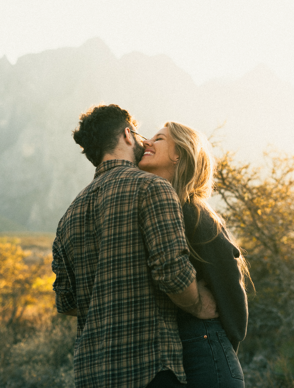 A romantic couple embraces outdoors during golden hour with mountains in the background.
