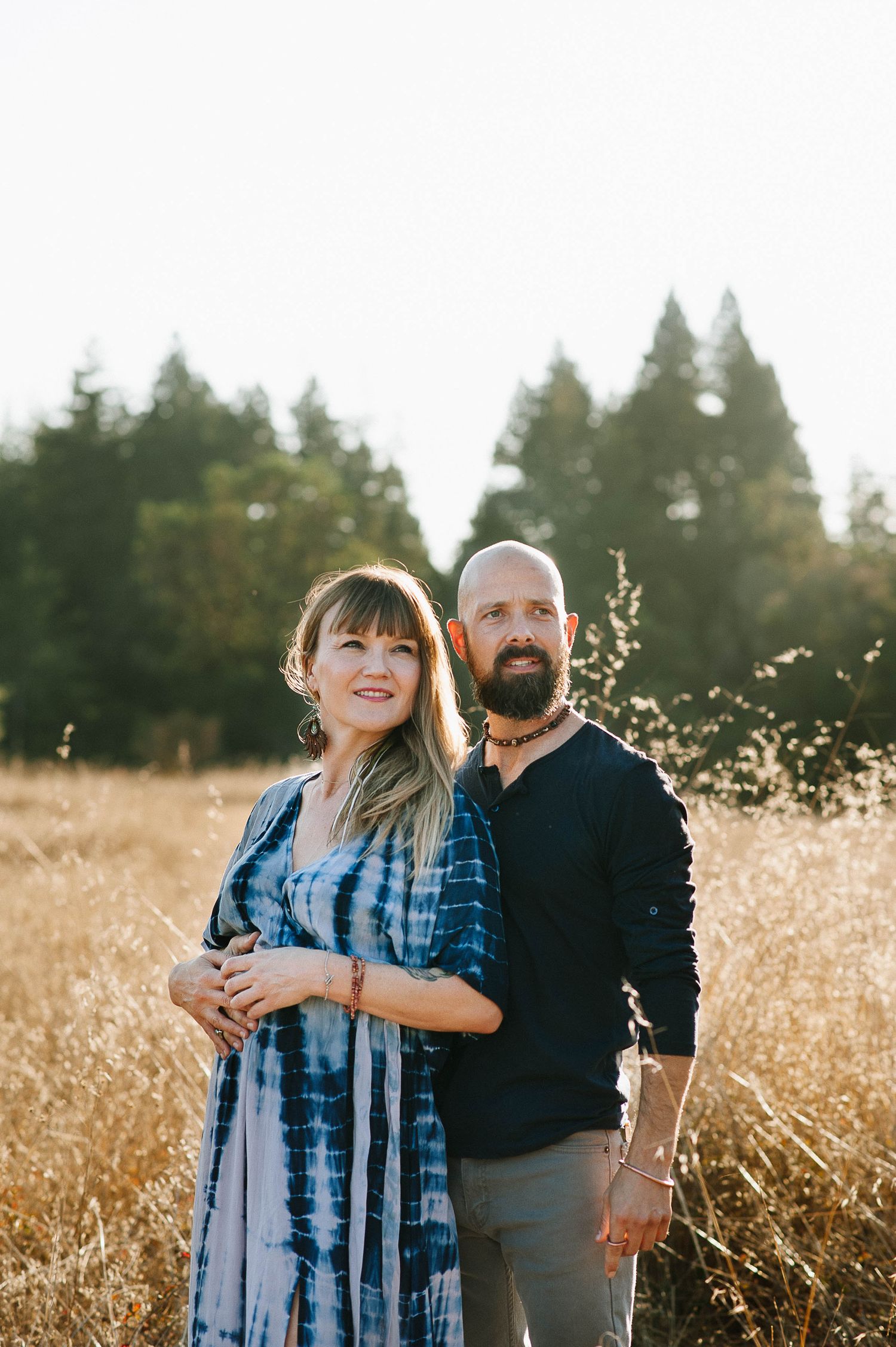 Couple standing together in sunlit meadow with evergreen forest in background.