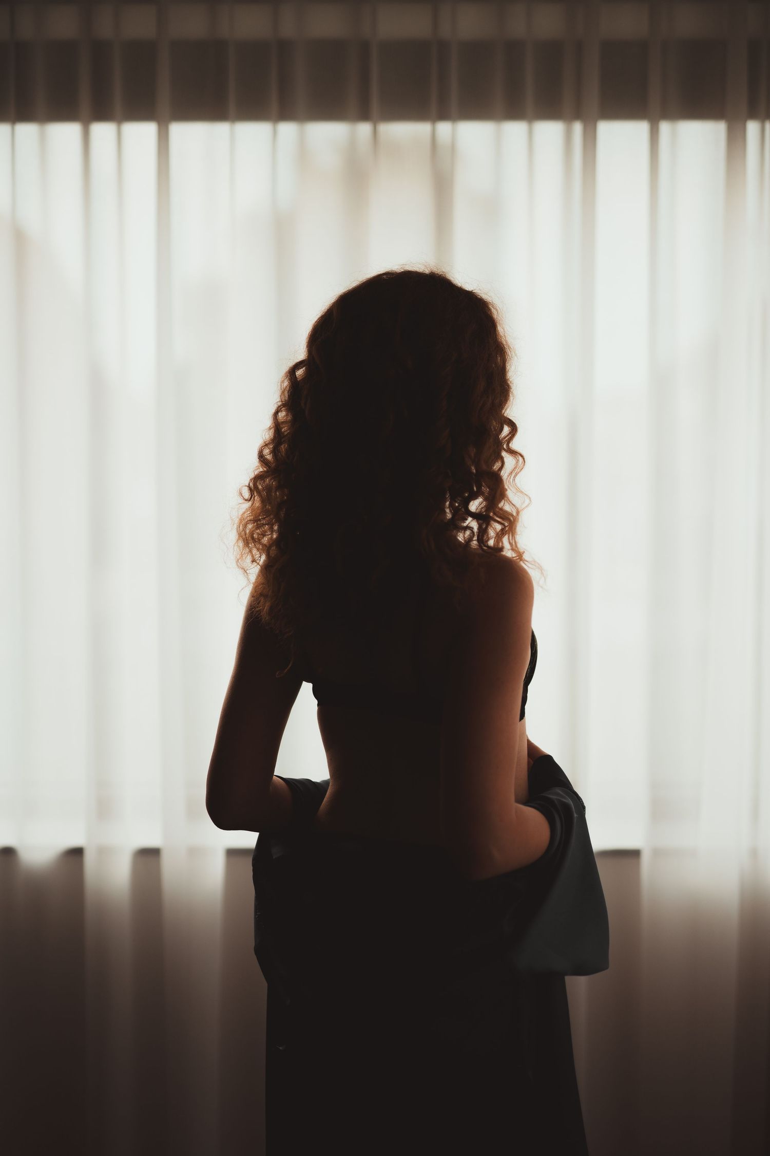 Femme rousse de dos debout devant une fenêtre lors de sa séance photo boudoir à Baudour près de Mons en Belgique