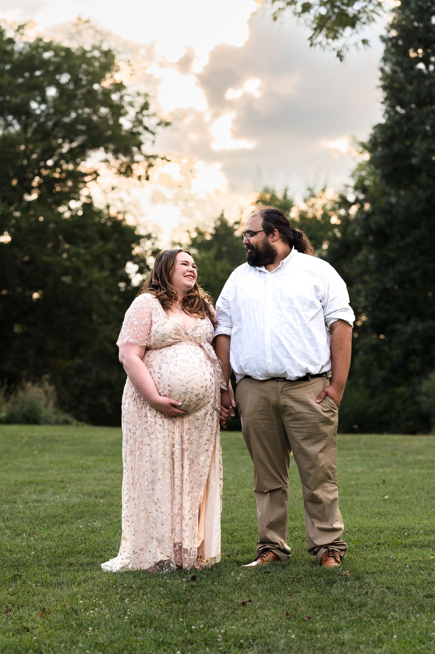 Expecting couple stands together on lawn during golden hour sunset.