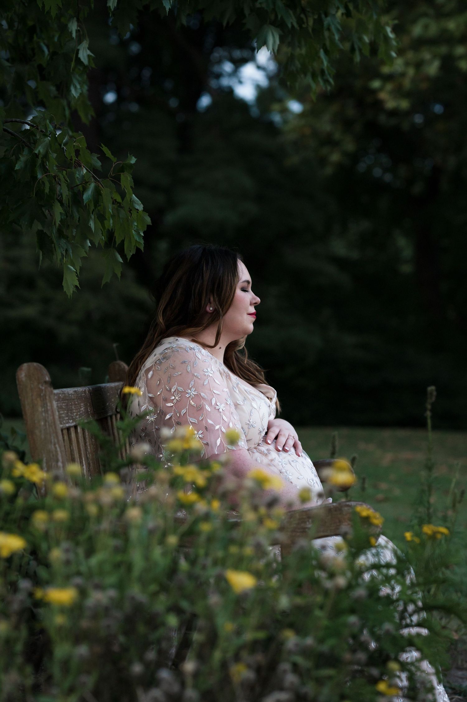 Pregnant woman sitting on wooden bench surrounded by garden flowers at dusk.