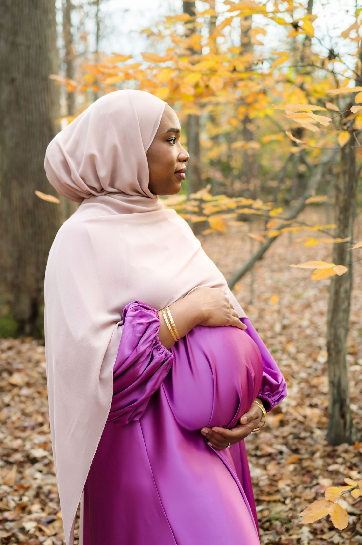 Woman in pink hijab and purple dress cradling her pregnant belly against autumn foliage.