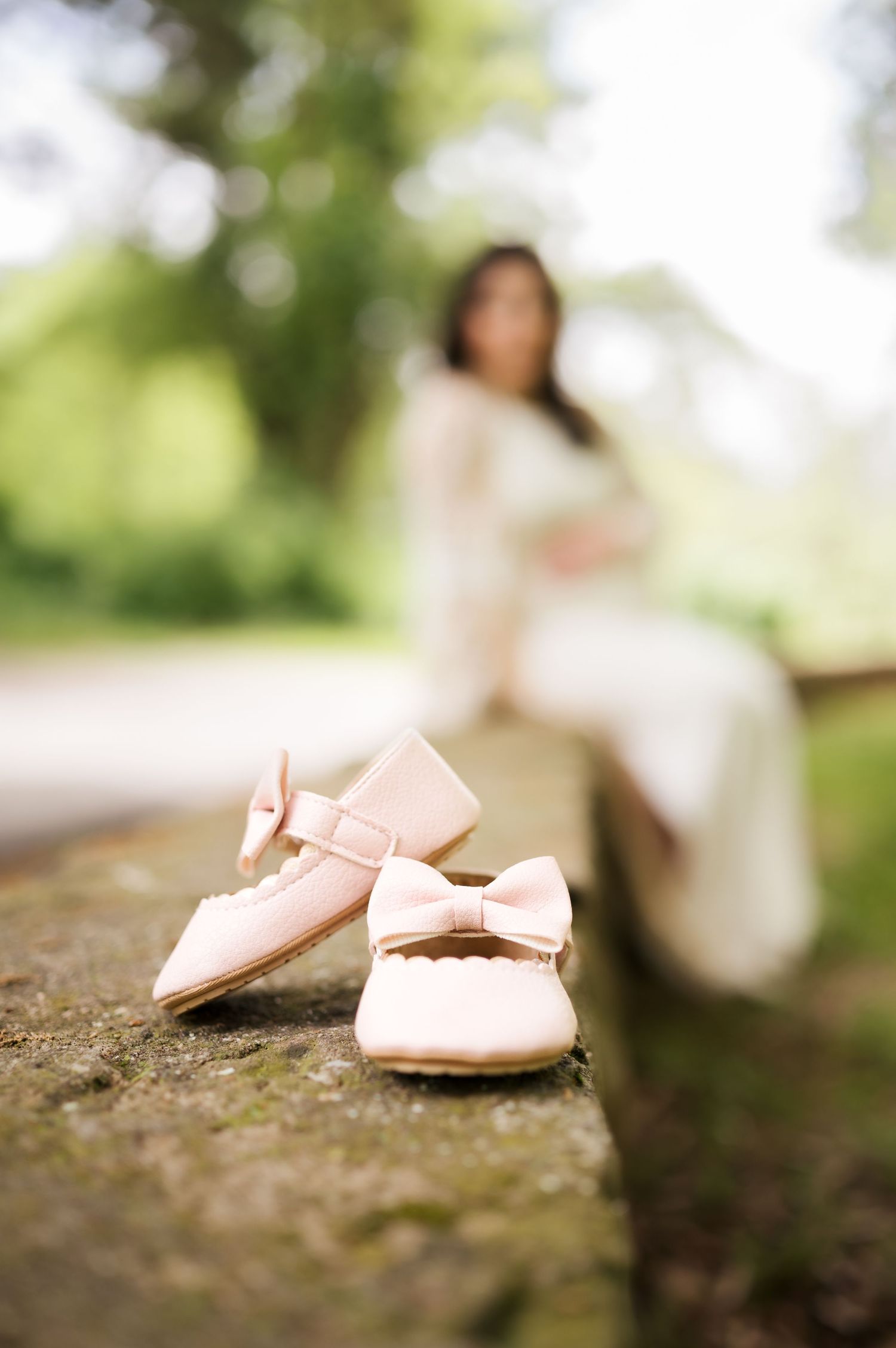 Pink baby shoes with bows sit in focus on stone with a blurred figure in white dress in background.