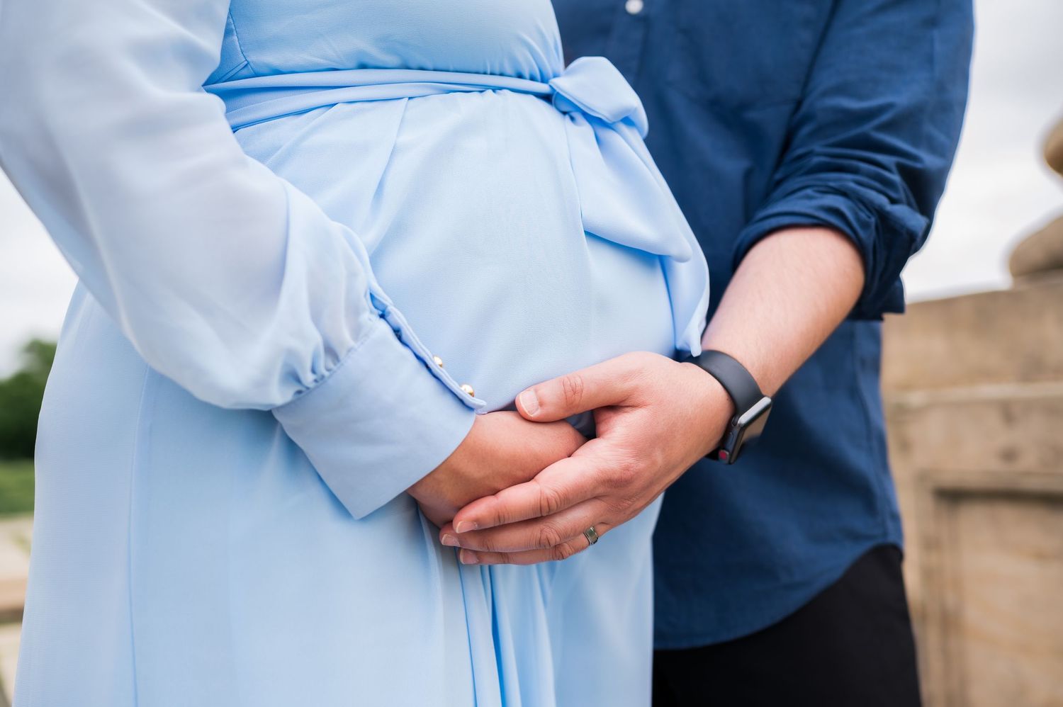 Side view of a pregnant woman in blue dress being embraced by partner.