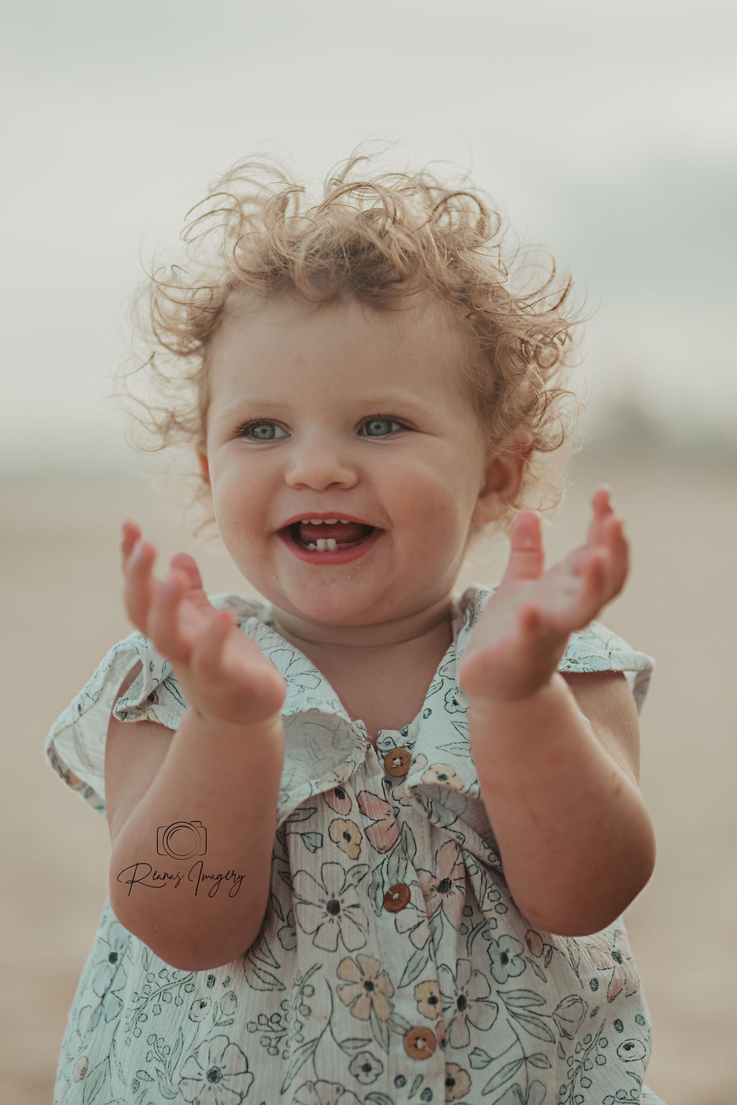 Curly-haired toddler girl smiling and clapping hands at the beach in a floral dress.