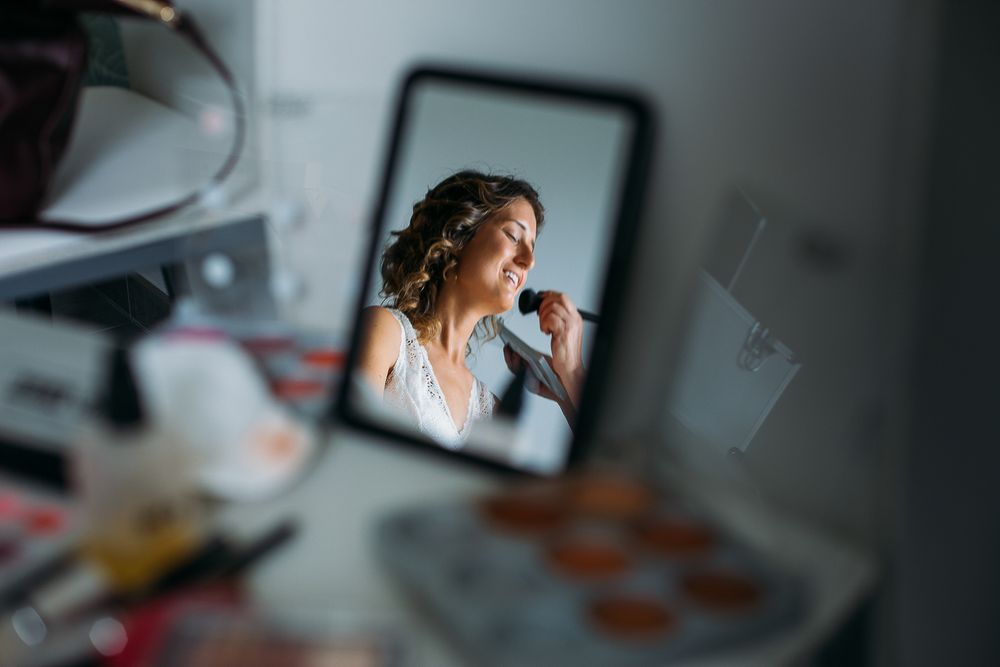 Mirror reflection shows bridal makeup preparation in soft natural light.