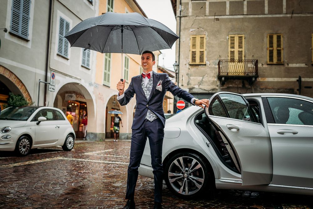 Stylish groom with umbrella poses in rain on European street near parked car.