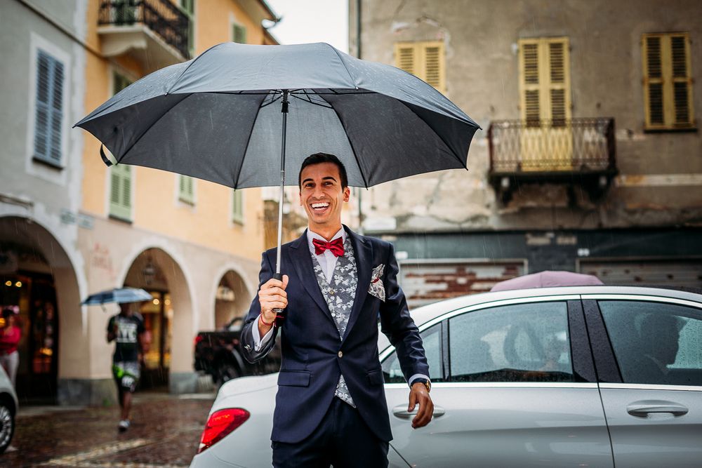 Groom holds large gray umbrella while standing in rainy European city street.