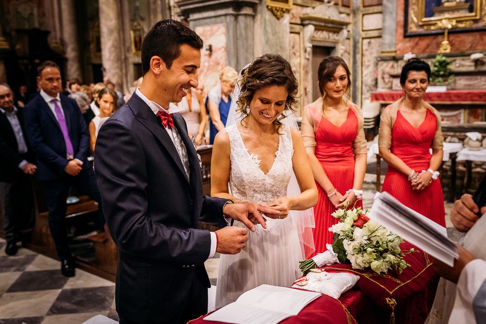 Couple signing marriage documents during traditional church ceremony with bridesmaids in red dresses looking on.