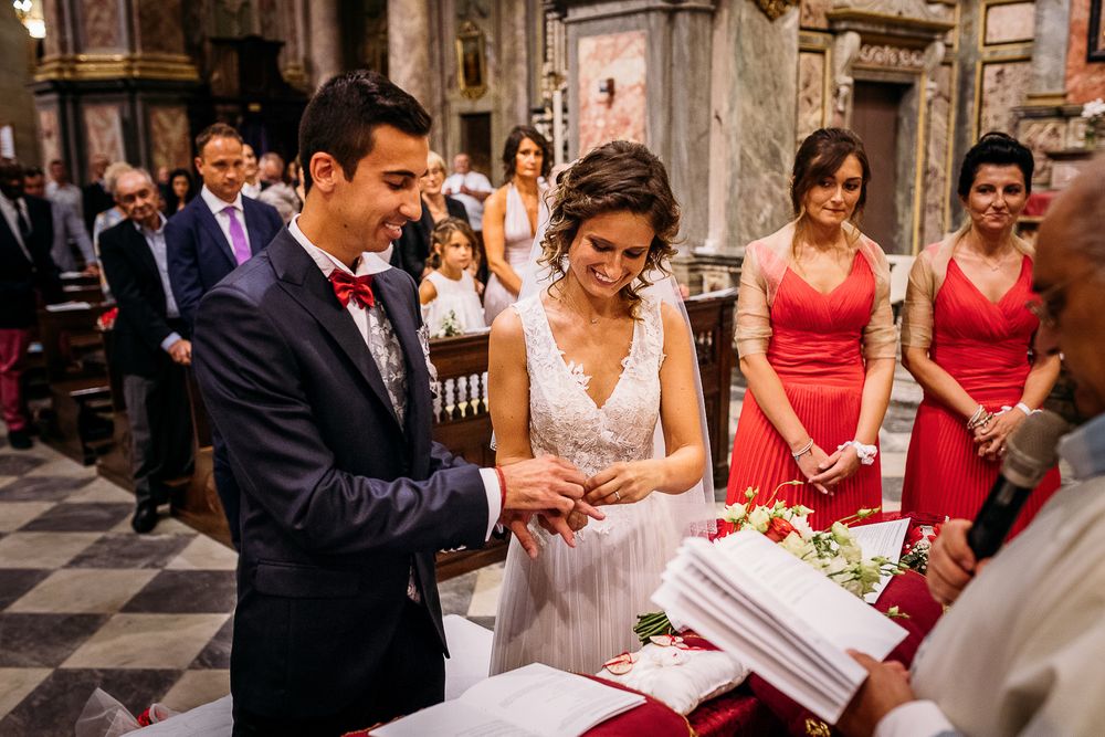 Wedding ceremony in ornate church as couple reviews documents with attendants in coral dresses nearby.