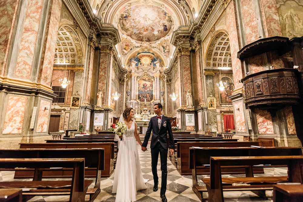 Newlyweds walking down aisle of ornately decorated baroque church with painted ceiling and wooden pews.
