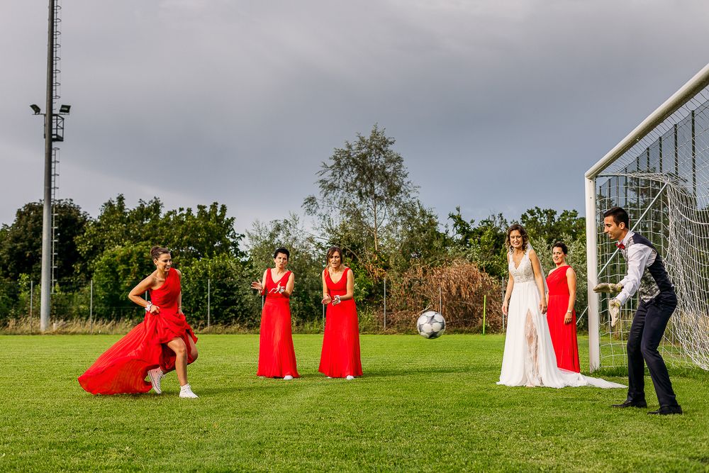 Wedding party in red dresses playing soccer on a green field under cloudy skies.