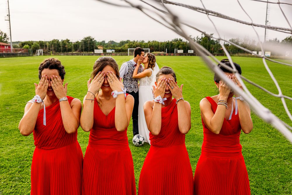 Bridesmaids in matching red dresses pose playfully covering their faces during outdoor photoshoot.