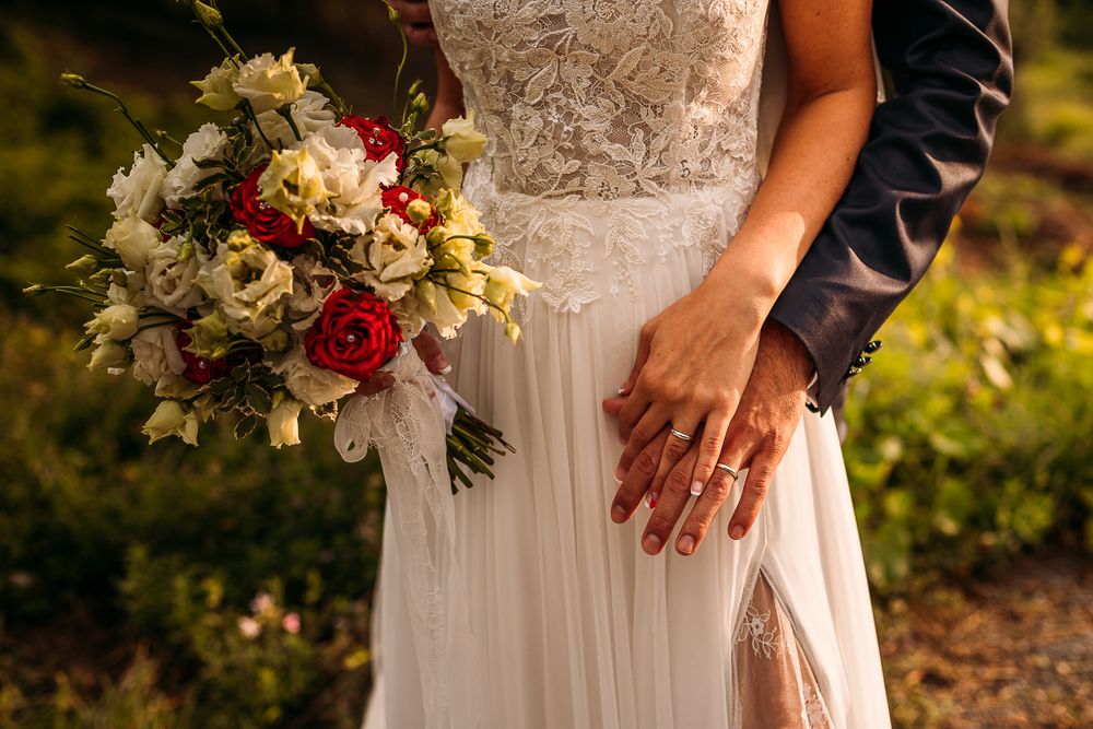 Wedding details showing white dress, hands with rings, and romantic red and white bouquet.