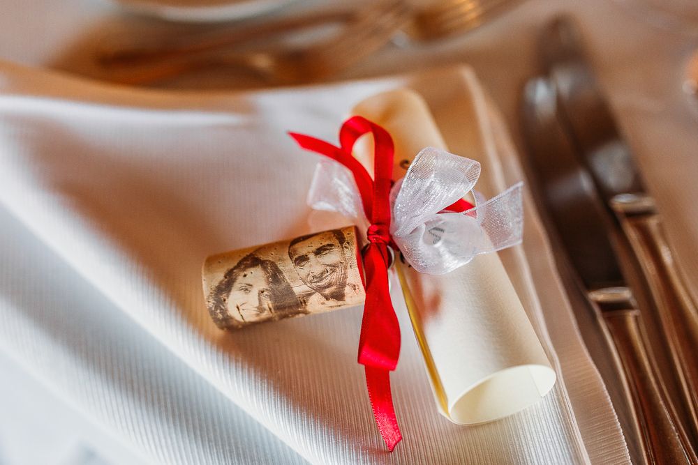 Wedding favor wrapped with red and white ribbons on white tablecloth next to silverware.