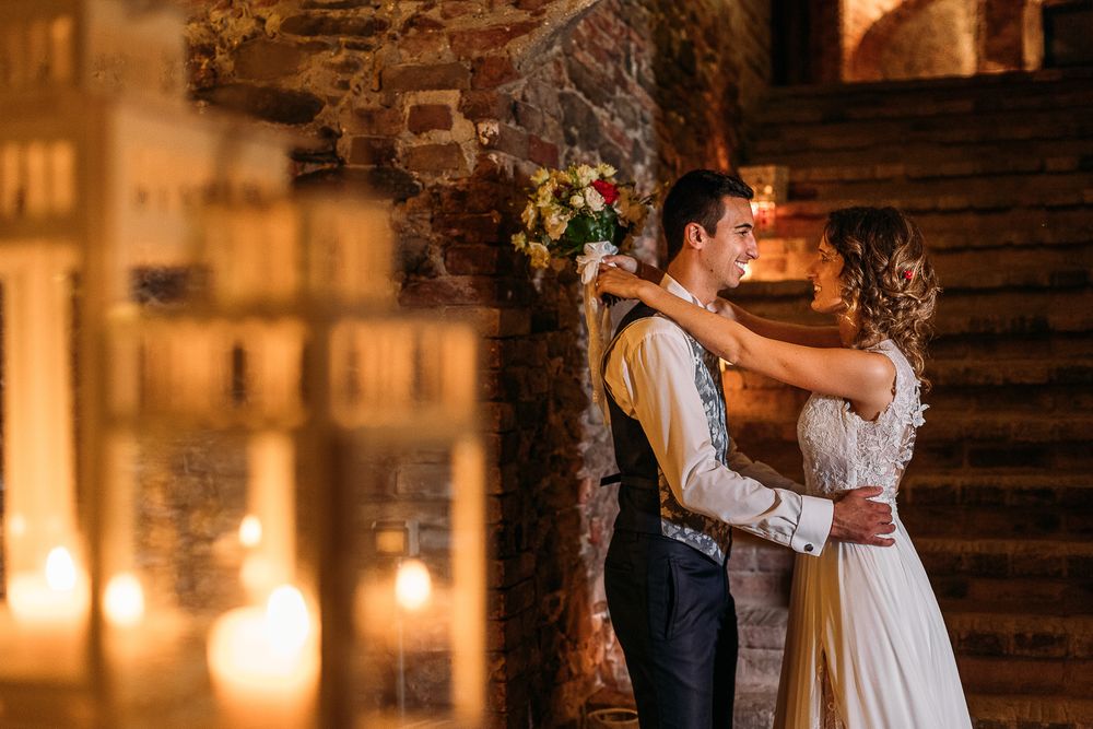 Romantic wedding scene with couple embracing in candlelit stone venue.