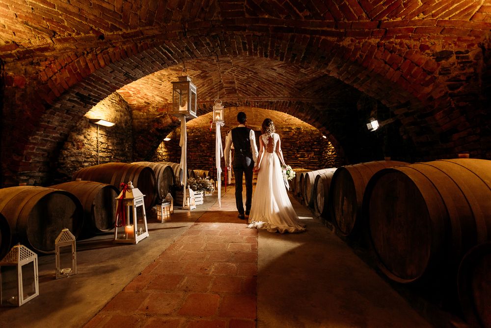 A wedding couple poses in a rustic wine cellar with wooden barrels and lantern lighting.