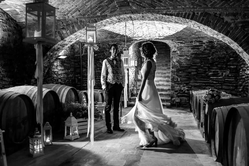 A black and white photograph of newlyweds in a wine barrel cellar with dramatic lighting.