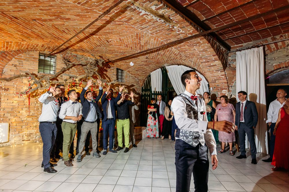 Wedding guests cheer and celebrate during a reception in a rustic brick-walled venue with string lights.