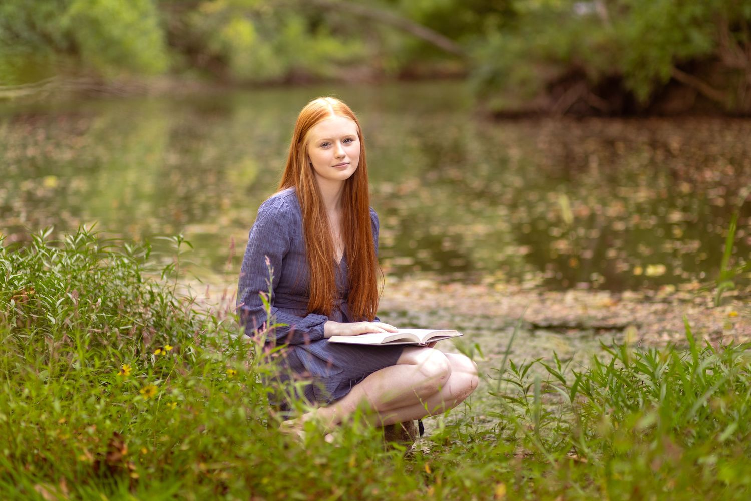 A high school senior girl sitting beside a stream, holding a book in her hands and smiling at the camera.