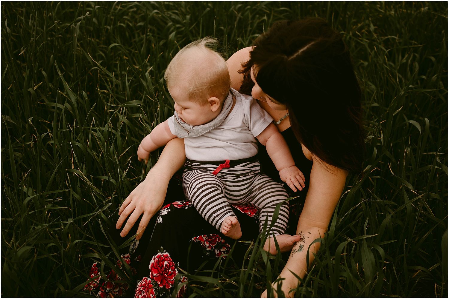 A baby in striped pants explores red flowers growing in a lush green field.