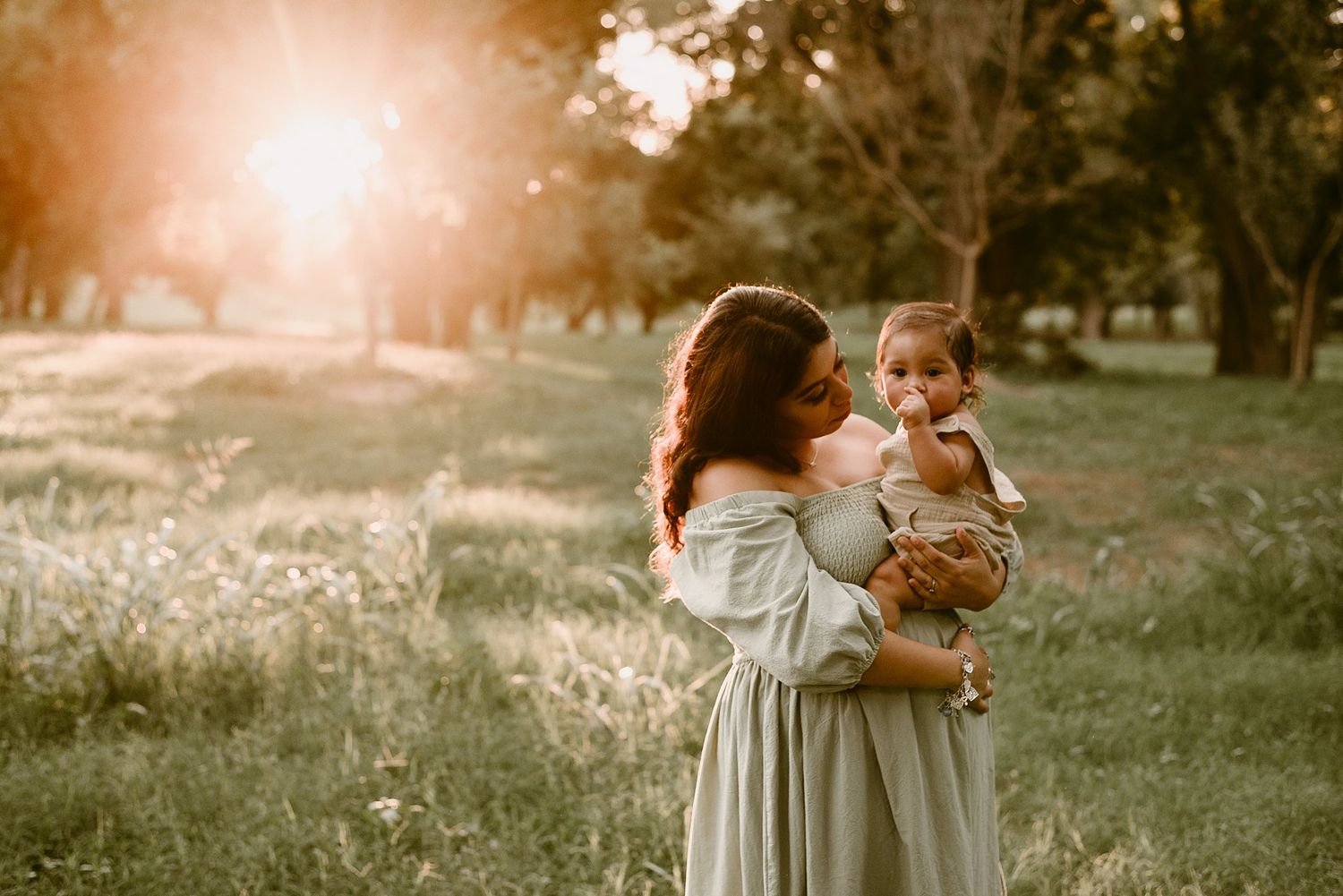 A woman in a light dress cradles a small child in a sun-drenched park setting.