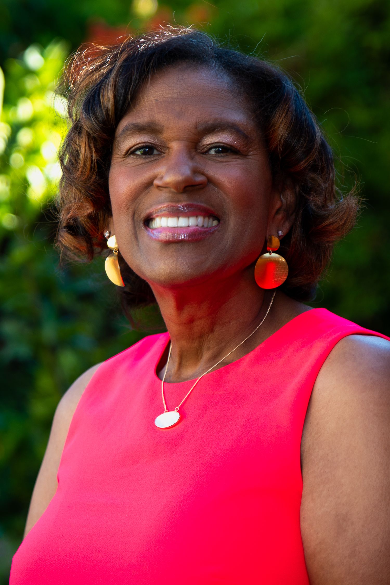 Portrait of someone in a vibrant red sleeveless top with gold jewelry against a green background.