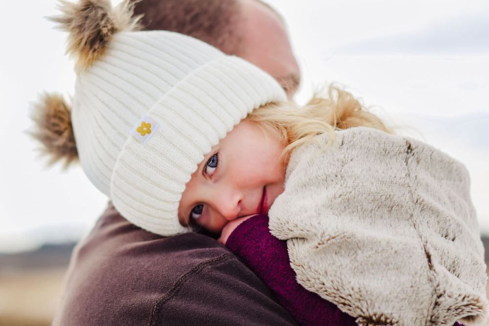 Blonde toddler in white pom pom hat snuggled against adult shoulder outdoors.