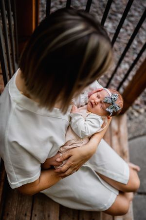 Parent holding newborn baby wearing white outfit and bow headband on wooden steps.