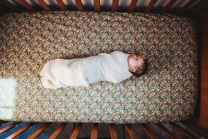 Newborn baby sleeping peacefully in white swaddle on floral patterned crib sheet.
