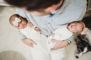 Twin newborns wearing white onesies being held in loving arms.