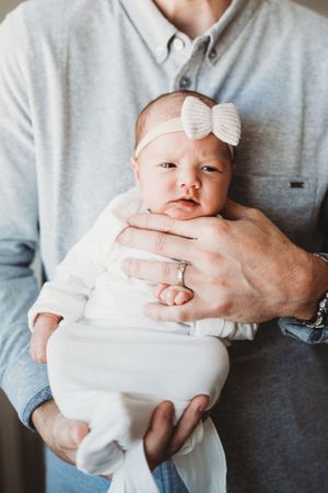 Newborn in white outfit and bow headband being cradled by person in light blue shirt.