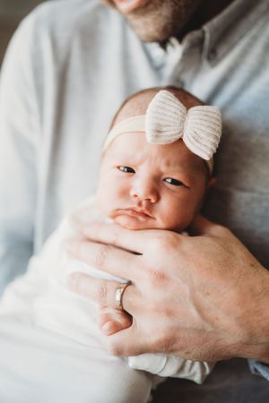 Baby wearing white bow headband held in arms with wedding ring visible.