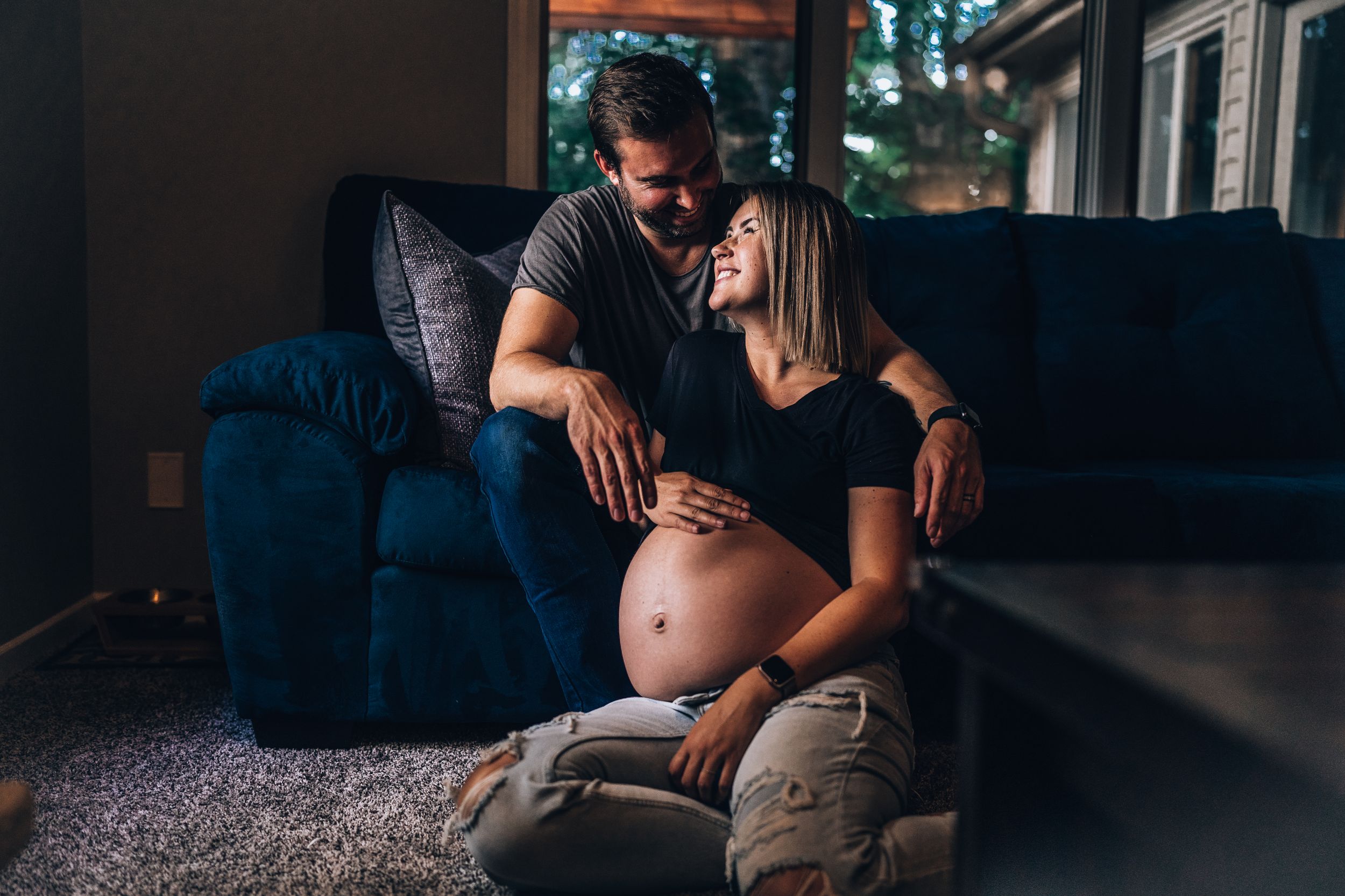 An intimate maternity moment captured on a blue couch in moody lighting.