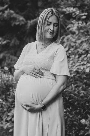 A pregnant person in a white dress poses outdoors in a black and white maternity photo.