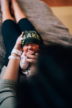 A baby's hand wearing hospital identification bands reaches toward a parent wearing a LAKE hat.