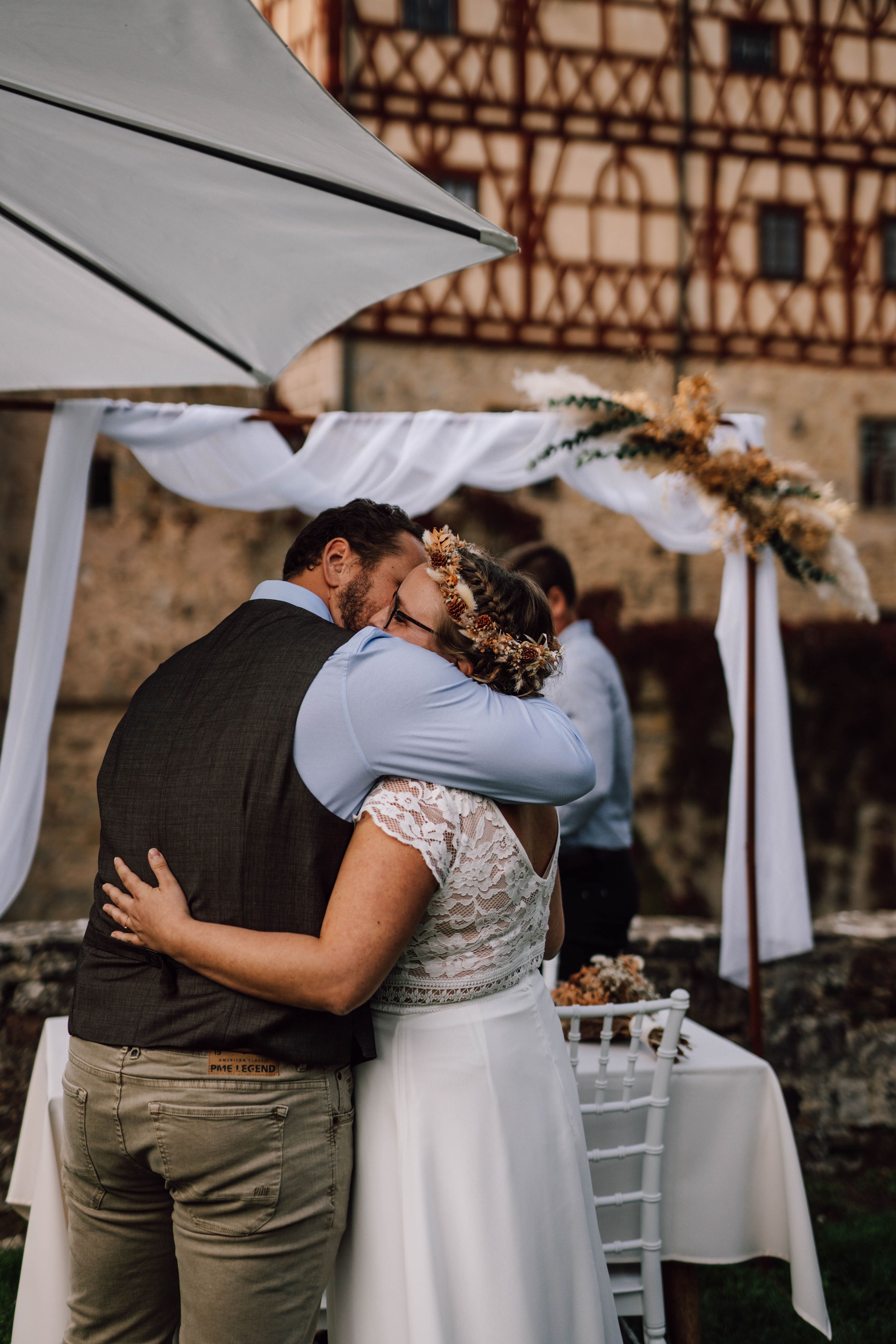 Wedding couple embracing under white umbrellas against traditional timber-frame building.