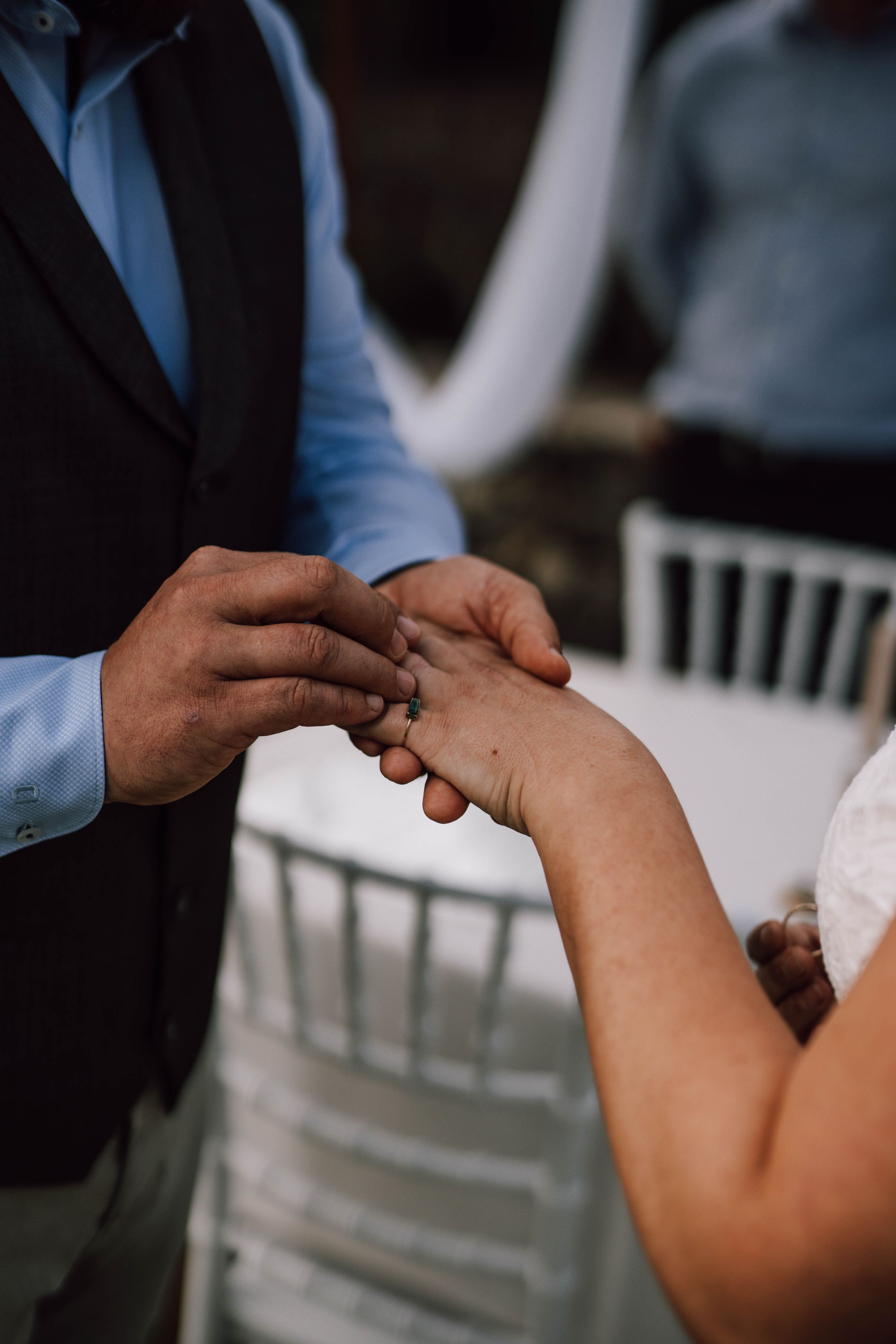 Close-up of hands exchanging rings during wedding ceremony with white chairs in background.