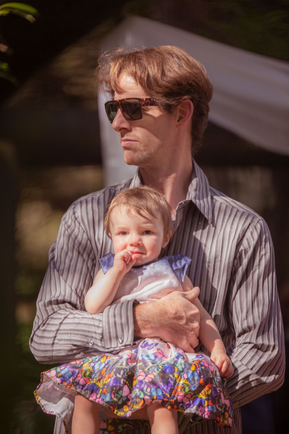 Guest in striped shirt and sunglasses holding young child in floral dress at outdoor event.