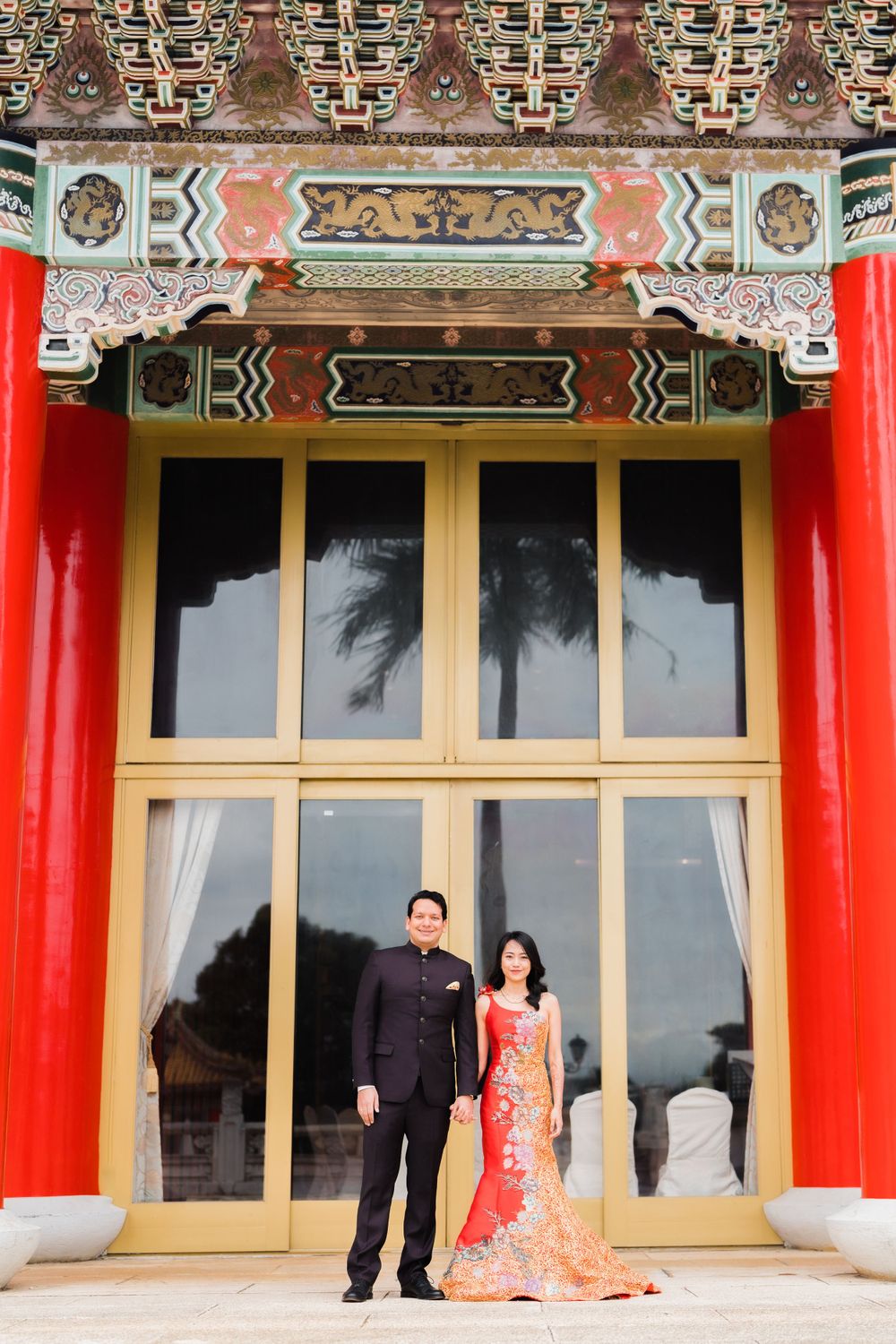 A couple stands formally in front of a traditional Asian building entrance with red columns and intricate details.