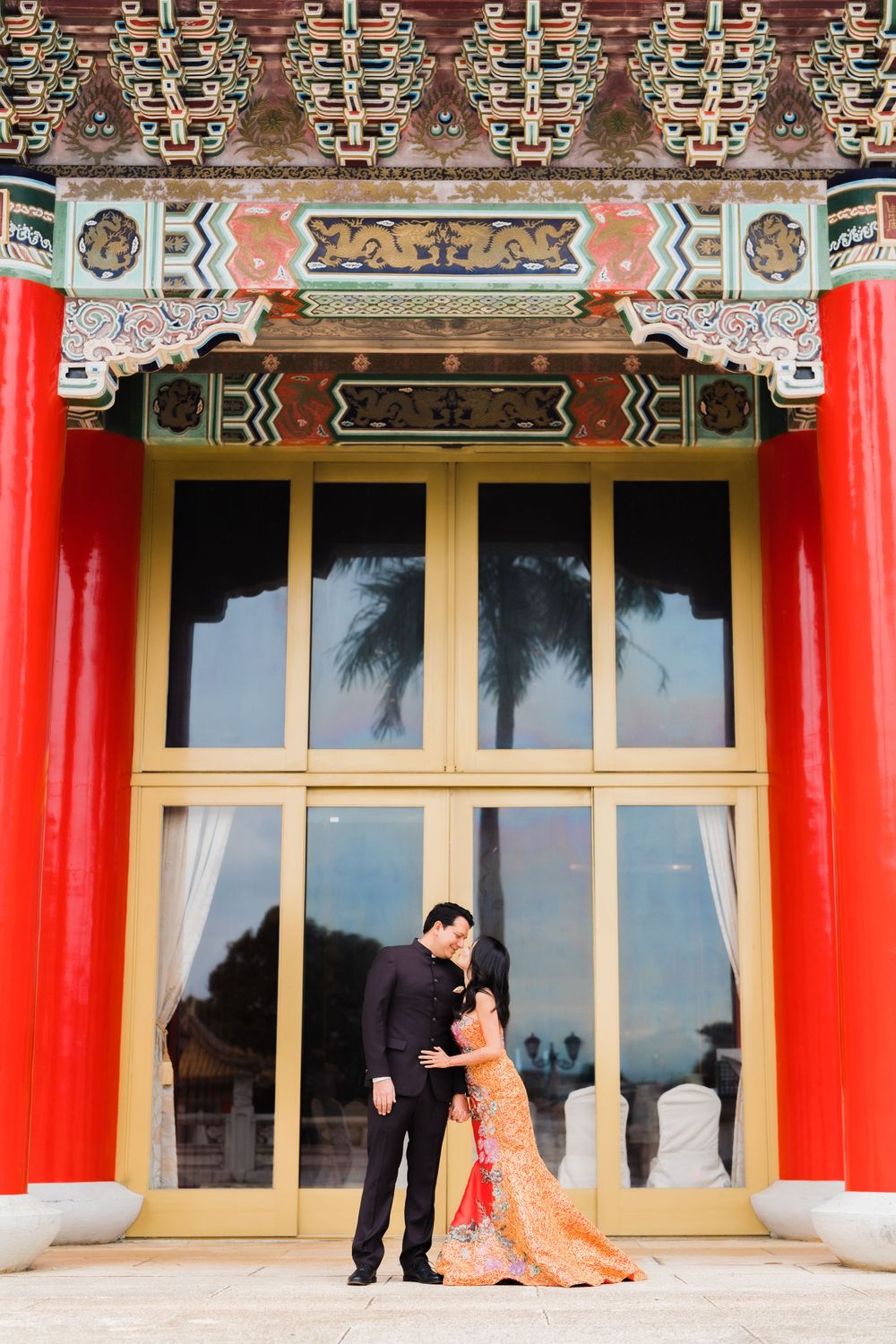 A couple poses romantically in front of an ornate Asian architectural entrance with red pillars and decorative details.