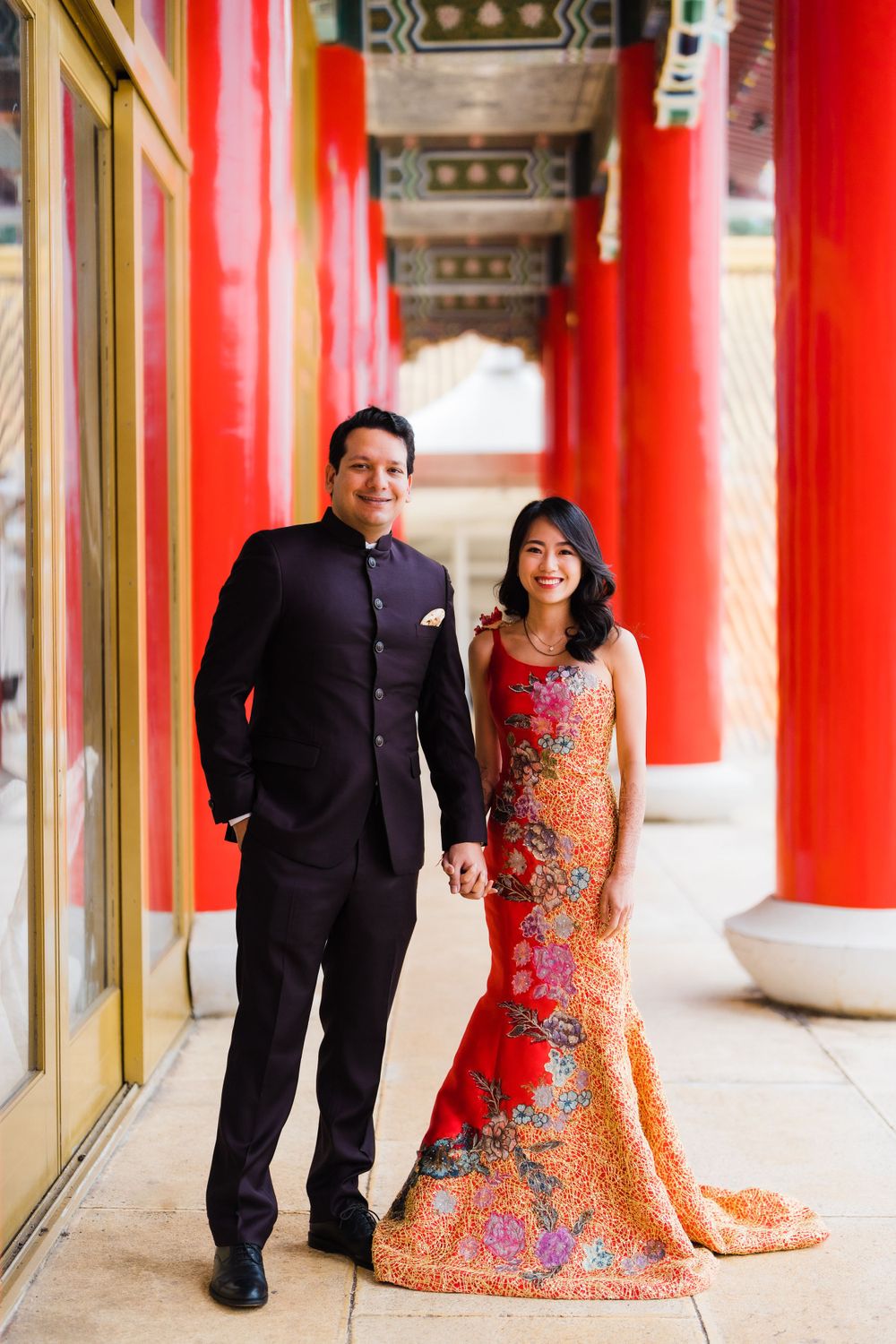 A couple poses in traditional formal wear against vibrant red pillars in an Asian-style corridor.