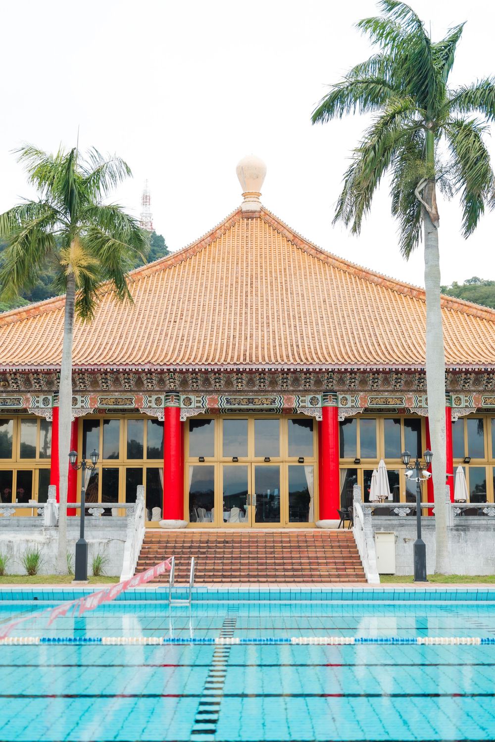 Traditional Chinese-style building with red pillars and tiled roof next to a swimming pool surrounded by palm trees.