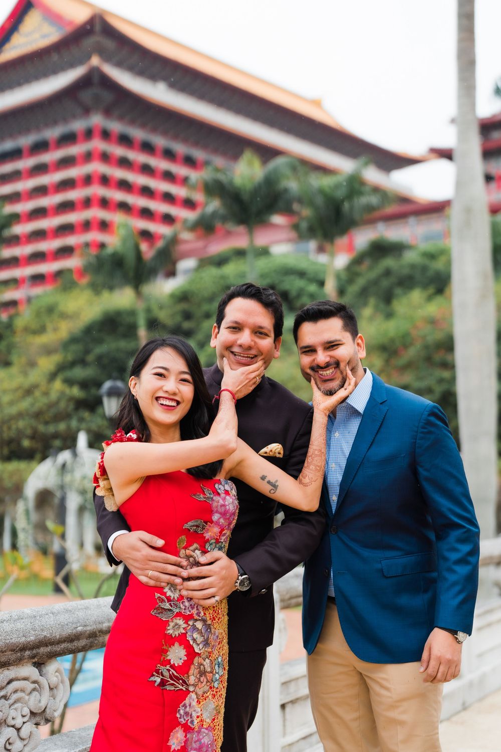 Friends pose playfully for photos outside a grand Asian architectural building with palm trees.