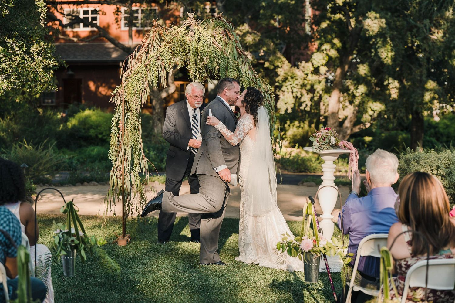 A garden wedding ceremony under a draped arch with white chairs and natural greenery.
