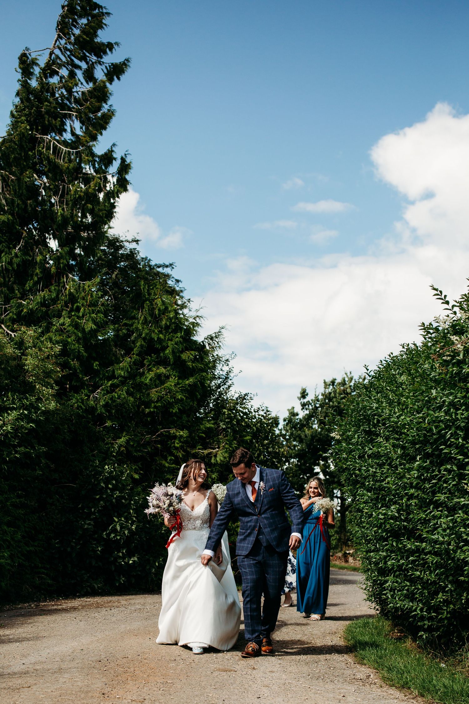 Wedding party walks along a tree-lined path under bright blue summer sky.