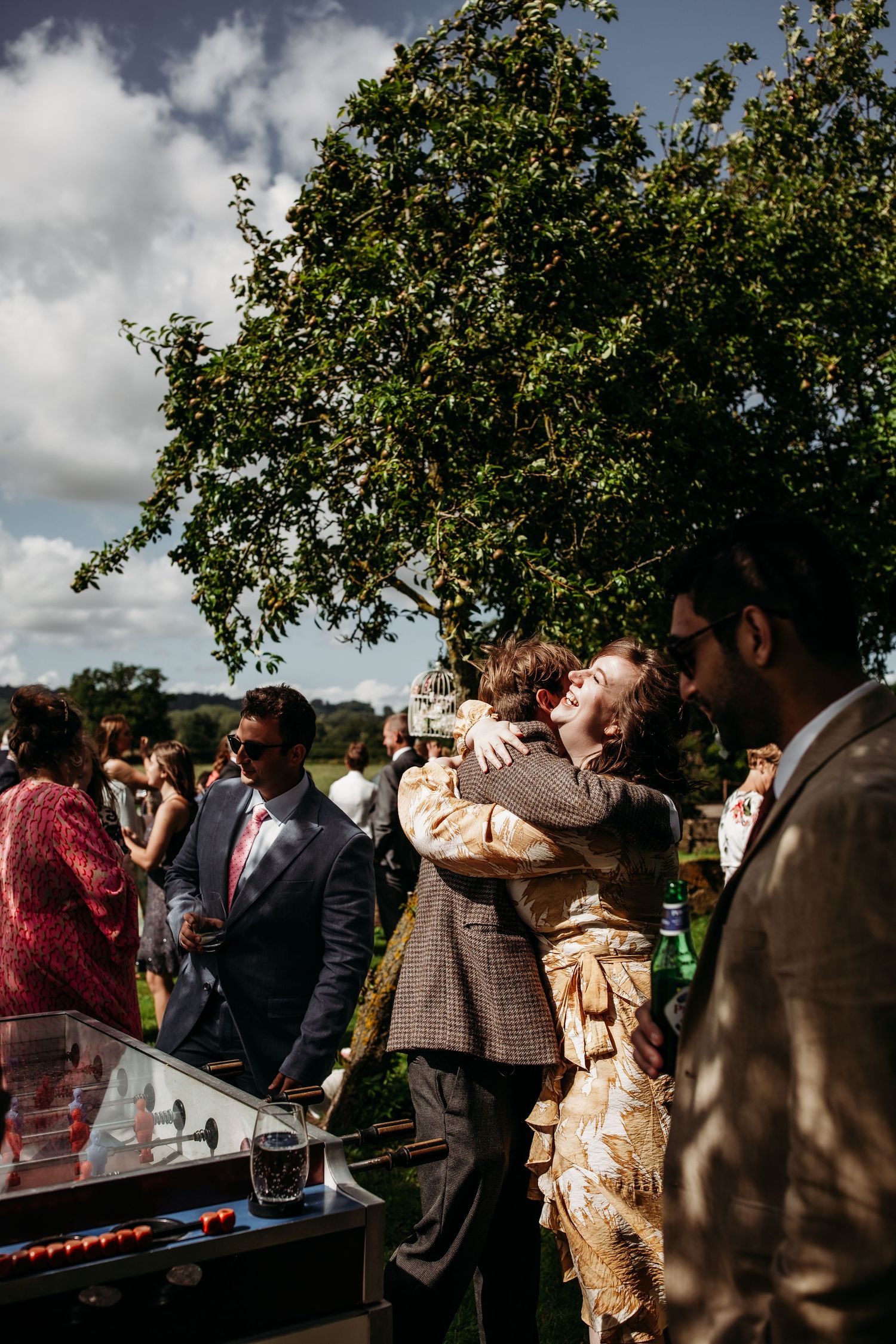 Outdoor celebration with guests gathered around entertainment equipment under leafy trees.
