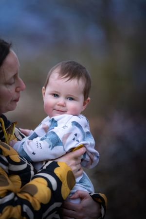 A baby in a white patterned sweater is held lovingly while smiling at the camera on a blurry background.
