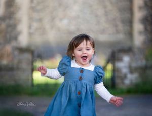A happy child wearing a blue dress with puffed sleeves laughs joyfully while playing outdoors.