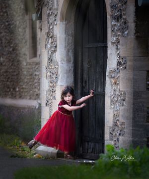 A young girl in a red velvet dress pushes open an old wooden door at a stone church entrance.